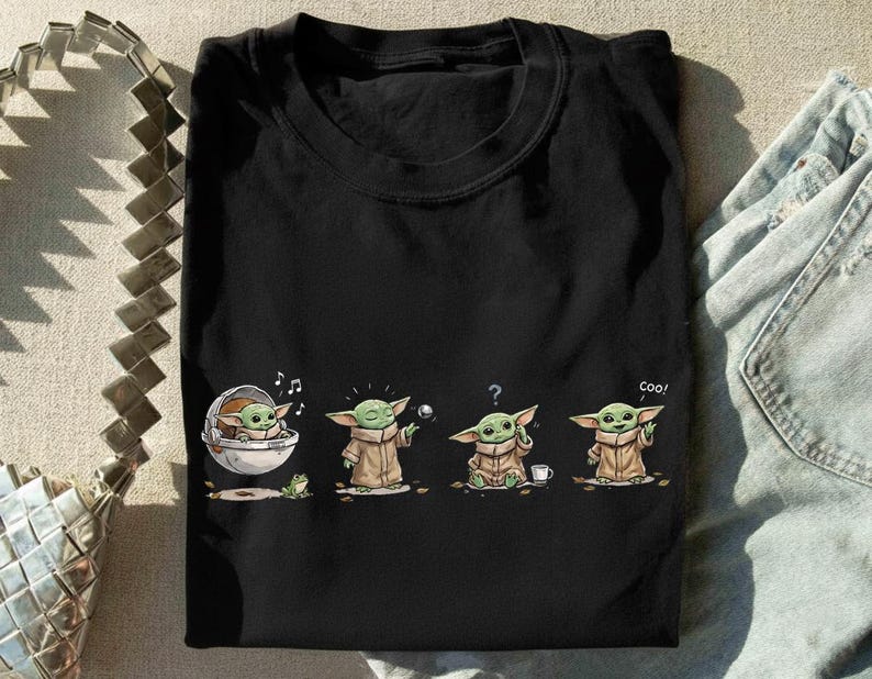 May include: A black short-sleeved t-shirt featuring a graphic print of a small green creature in a tan robe. The illustration shows the character in four poses: sitting in a floating pod, reaching for a ball, looking confused, and saying coo.