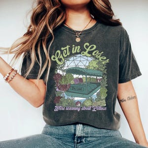 May include: Dark gray t-shirt with the text "Get in Loser" in green script above a graphic of a boat surrounded by plants. The text "The Land" is on the boat, and "We're learning about Lettuce" is below.