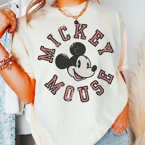 May include: Cream-colored t-shirt featuring a distressed graphic of Mickey Mouse with the text "MICKEY MOUSE" in a circular design. The design is in black and pink. The shirt has short sleeves and a relaxed fit.