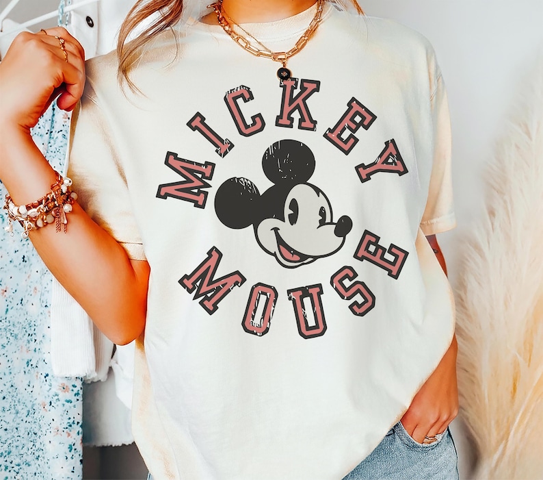 May include: Cream-colored t-shirt featuring a distressed graphic of Mickey Mouse with the text "MICKEY MOUSE" in a circular design. The design is in black and pink. The shirt has short sleeves and a relaxed fit.
