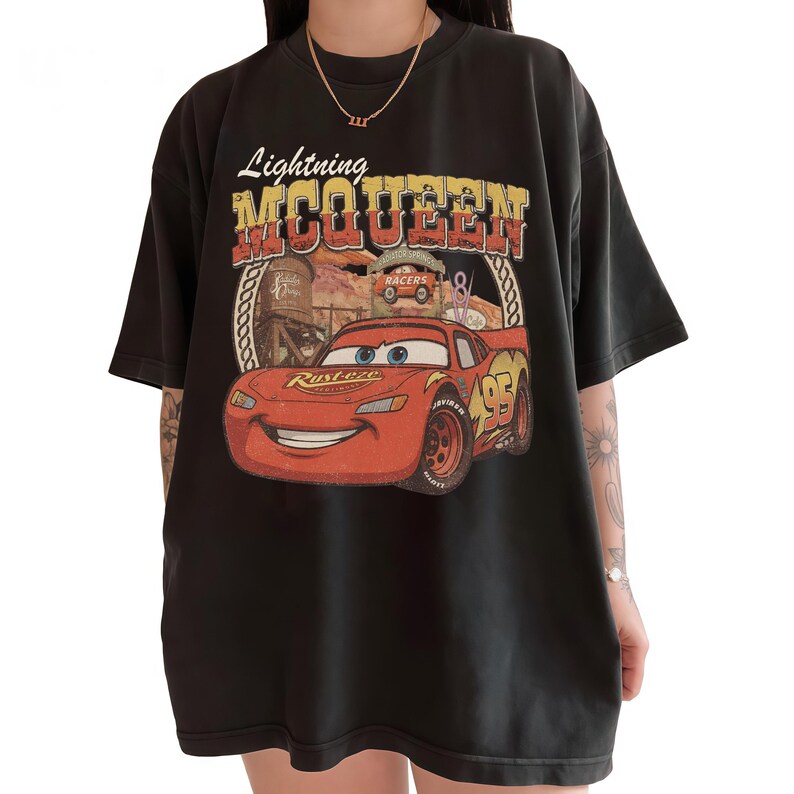 Puede incluir: Una camiseta negra de manga corta con un estampado gr&aacute;fico del personaje Lightning McQueen. El dise&ntilde;o incluye el texto "Lightning MCQUEEN" en una fuente desgastada, un fondo de paisaje des&eacute;rtico y el n&uacute;mero 95 en el lateral del coche.