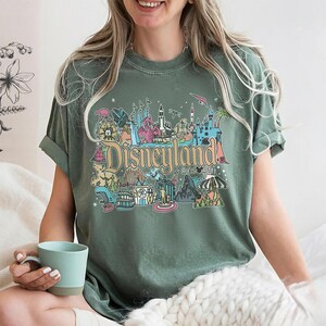 May include: Green t-shirt with a colorful Disneyland Resort graphic. The design features iconic park imagery and the Disneyland Resort text in a vintage style. The shirt has short sleeves and a relaxed fit.