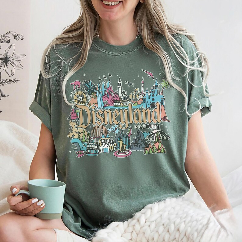 May include: Green t-shirt with a colorful Disneyland Resort graphic. The design features iconic park imagery and the Disneyland Resort text in a vintage style. The shirt has short sleeves and a relaxed fit.