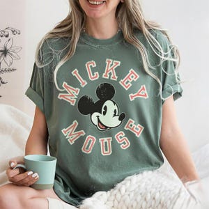 May include: Sage green t-shirt featuring a graphic of Mickey Mouse with the words "Mickey Mouse" in a circular design. The design is in red and white. The shirt has short sleeves and a relaxed fit.
