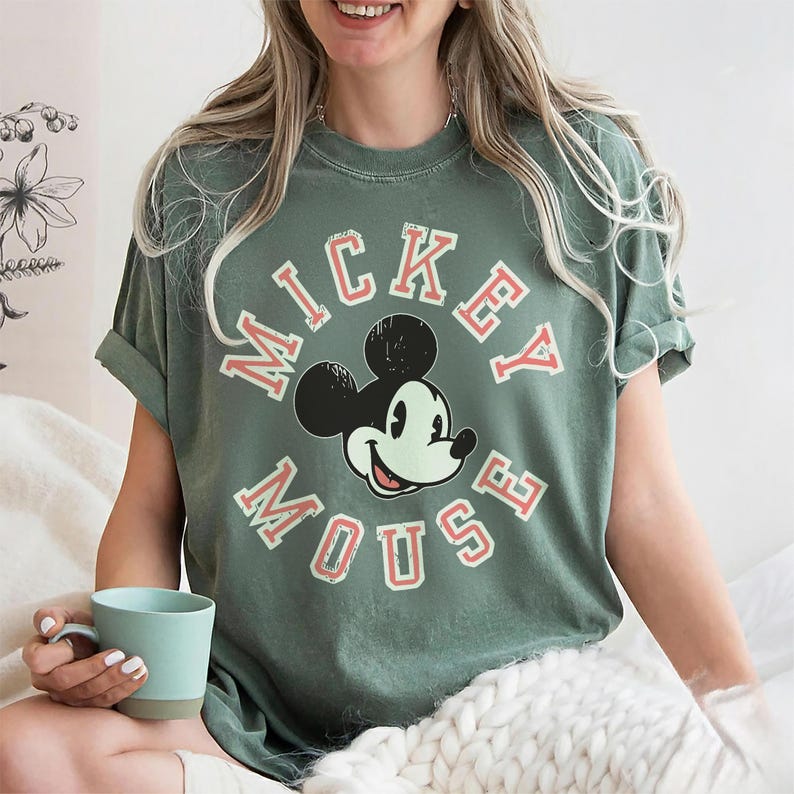 May include: Sage green t-shirt featuring a graphic of Mickey Mouse with the words "Mickey Mouse" in a circular design. The design is in red and white. The shirt has short sleeves and a relaxed fit.