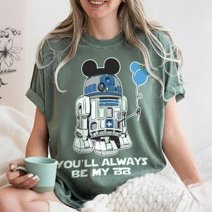 May include: Green t-shirt featuring a cartoon R2-D2 character with Mickey Mouse ears and a blue balloon. The text "YOU'LL ALWAYS BE MY BB" is printed below the graphic. The shirt is a relaxed fit.