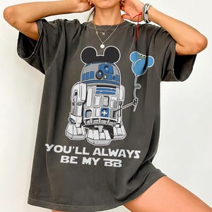 May include: Dark grey t-shirt featuring a cartoon R2-D2 character with Mickey Mouse ears holding a blue flower balloon. The text "YOU'LL ALWAYS BE MY BB" is printed in white below the graphic.