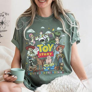 May include: Green t-shirt featuring a colorful graphic of characters from the animated film "Toy Story." The design includes Woody, Buzz Lightyear, and other characters, with the words "TOY STORY" and "YOU'VE GOT A FRIEND IN ME" printed on the front. The shirt has short sleeves.