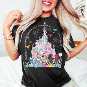May include: A black t-shirt with a graphic print of Cinderella, Snow White, Ariel, Belle, Rapunzel, and Aurora standing in front of Cinderella's castle with fireworks in the background.