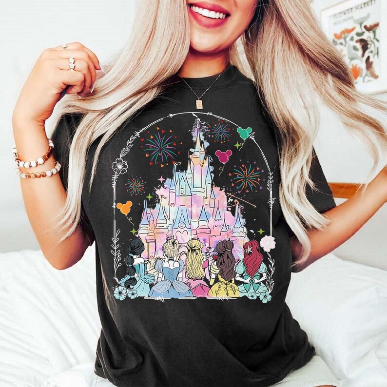 May include: A black t-shirt with a graphic print of Cinderella, Snow White, Ariel, Belle, Rapunzel, and Aurora standing in front of Cinderella's castle with fireworks in the background.
