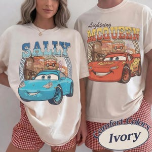 Puede incluir: Dos camisetas de color blanco roto con estampados gr&aacute;ficos de la pel&iacute;cula Cars. La camiseta de la izquierda muestra un coche azul con el texto SALLY, mientras que la de la derecha muestra un coche rojo con el texto Lightning MCQUEEN.