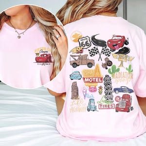 May include: Light pink t-shirt featuring a graphic of a red race car with the text "Piston Cup Champion '95". The back of the shirt has various car-themed illustrations, including Route 66 signage and checkered flags.