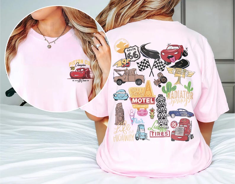 May include: Light pink t-shirt featuring a graphic of a red race car with the text "Piston Cup Champion '95". The back of the shirt has various car-themed illustrations, including Route 66 signage and checkered flags.