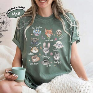 May include: A moss green t-shirt featuring a graphic print of various animated animal characters including a fox, rabbit, sheep, and sloth. Each character is labeled with names such as Nick Wilde, Judy Hopps, and Flash, surrounded by small stars.