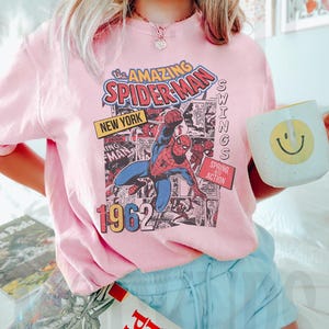 Peut inclure: T-shirt rose clair avec un imprim&eacute; de bande dessin&eacute;e Spider-Man. Le graphisme comprend le texte "the AMAZING SPIDER-MAN NEW YORK 1962" et "SPRING INTO ACTION". Une tasse blanche avec un smiley jaune est tenue dans l'image.