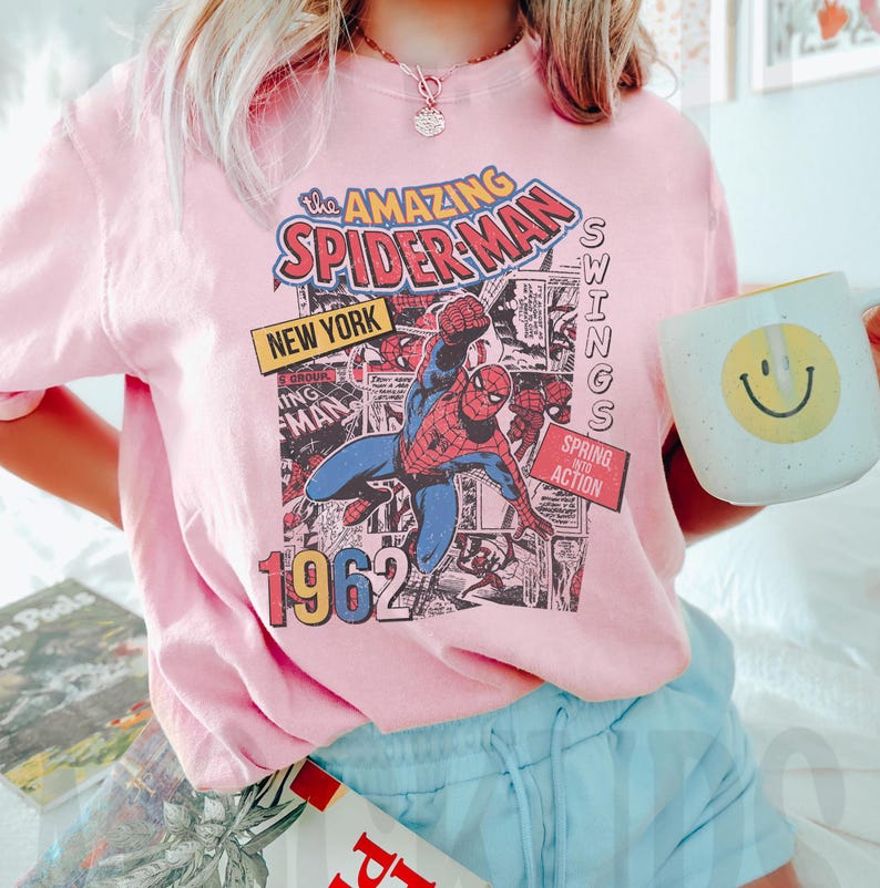 Peut inclure: T-shirt rose clair avec un imprim&eacute; de bande dessin&eacute;e Spider-Man. Le graphisme comprend le texte "the AMAZING SPIDER-MAN NEW YORK 1962" et "SPRING INTO ACTION". Une tasse blanche avec un smiley jaune est tenue dans l'image.