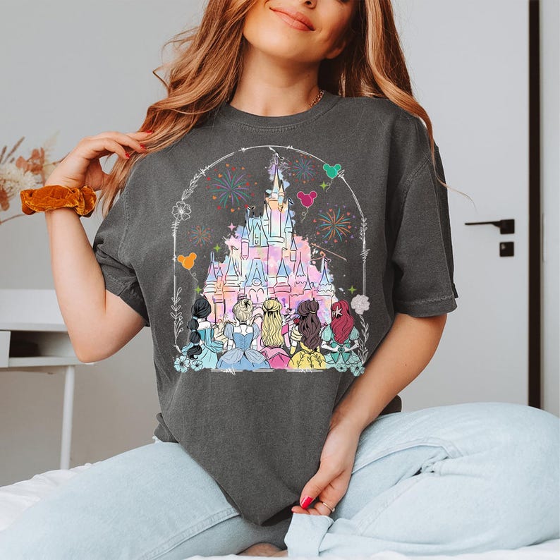 May include: A charcoal grey t-shirt with a graphic of Cinderella's castle and Disney princesses. The princesses are standing in front of the castle, which is surrounded by fireworks.
