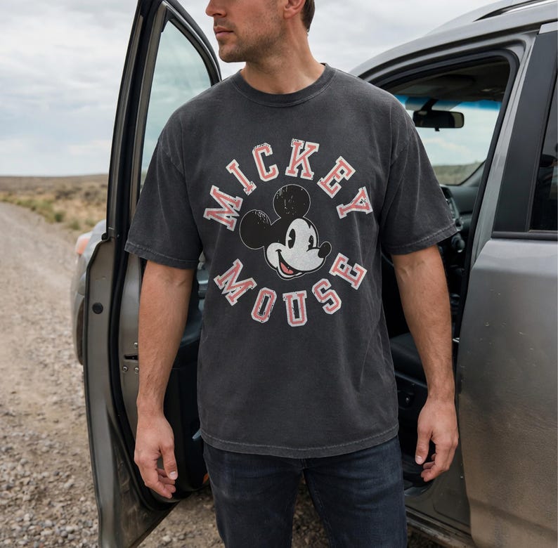 May include: A charcoal gray t-shirt featuring a distressed Mickey Mouse graphic. The design includes the words "Mickey Mouse" in a circular arrangement with red accents. The shirt is paired with dark blue jeans.