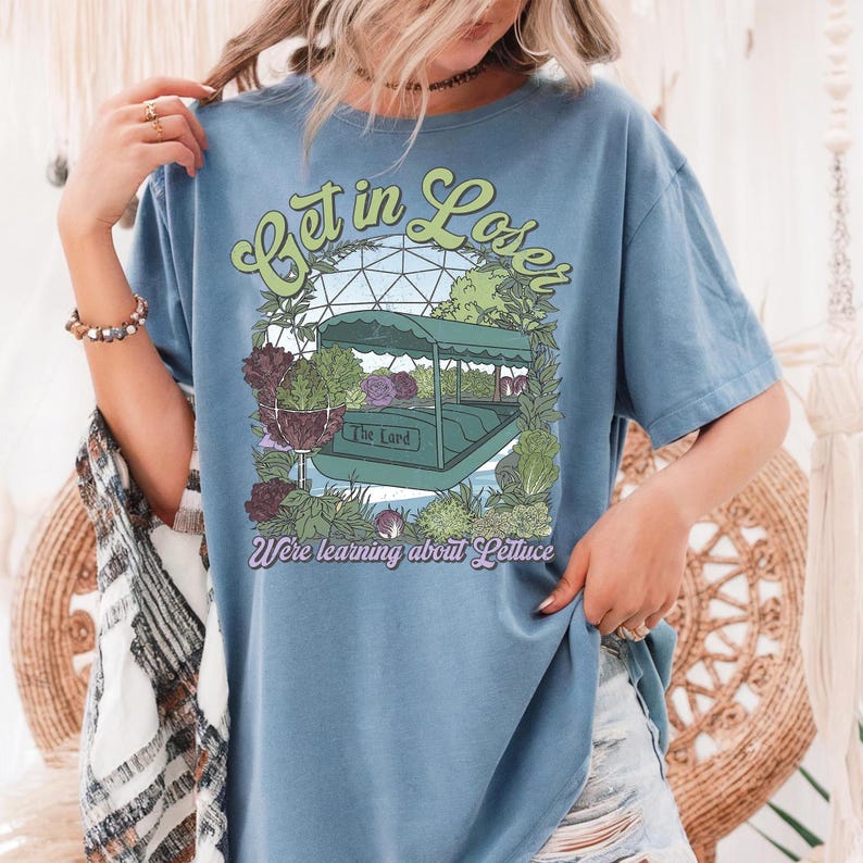 May include: Blue t-shirt with a graphic featuring a boat ride through a greenhouse with the text "Get in Loser" and "We're learning about Lettuce". The design includes lettuce and other plants.