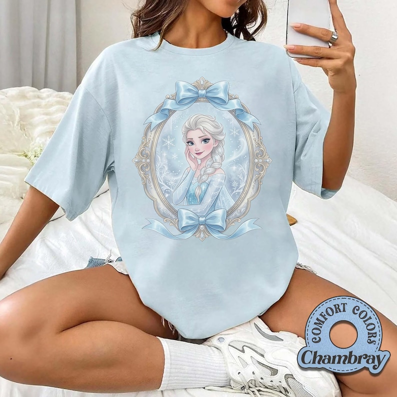 K&ouml;nnte beinhalten: Ein hellblaues T-Shirt mit einem Grafikdruck einer blonden Figur mit geflochtener Frisur in einem verzierten, ovalen Rahmen, der mit hellblauen Schleifen und Schneeflocken verziert ist. Das Motiv ist mittig auf der Vorderseite platziert.