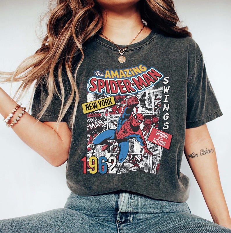 Peut inclure: T-shirt gris fonc&eacute; avec un design de bande dessin&eacute;e vintage de Spider-Man. Le graphisme comprend le texte "The Amazing Spider-Man", "New York" et "1962". Le super-h&eacute;ros est repr&eacute;sent&eacute; dans son costume rouge et bleu classique.
