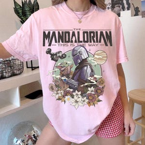 Puede incluir: Una camiseta de manga corta de color rosa claro con un estampado gr&aacute;fico de The Mandalorian y Grogu. El texto dice THE MANDALORIAN THIS IS THE WAY. El dise&ntilde;o incluye una nave espacial, un planeta y un grupo de flores coloridas en la parte inferior.