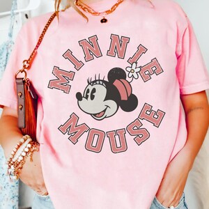 Puede incluir: Camiseta rosa claro con un dise&ntilde;o de Minnie Mouse y el texto "MINNIE MOUSE" en un dise&ntilde;o circular. Minnie Mouse tiene una cabeza negra, un lazo rosa y una flor blanca. La camiseta es de color rosa claro.