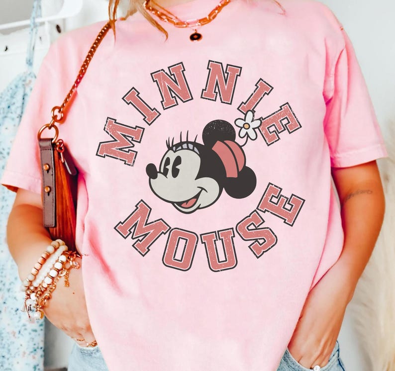 Puede incluir: Camiseta rosa claro con un dise&ntilde;o de Minnie Mouse y el texto "MINNIE MOUSE" en un dise&ntilde;o circular. Minnie Mouse tiene una cabeza negra, un lazo rosa y una flor blanca. La camiseta es de color rosa claro.