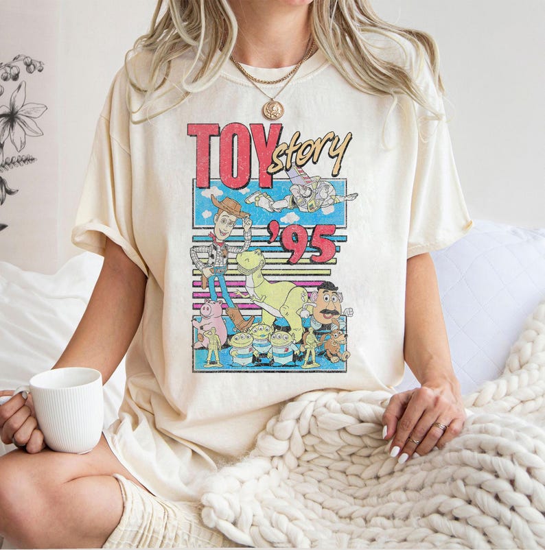 May include: Cream-colored t-shirt featuring a colorful graphic from the movie "Toy Story." The design includes the text "Toy Story '95" with images of Woody, Buzz Lightyear, and other characters. The shirt has short sleeves and a relaxed fit.