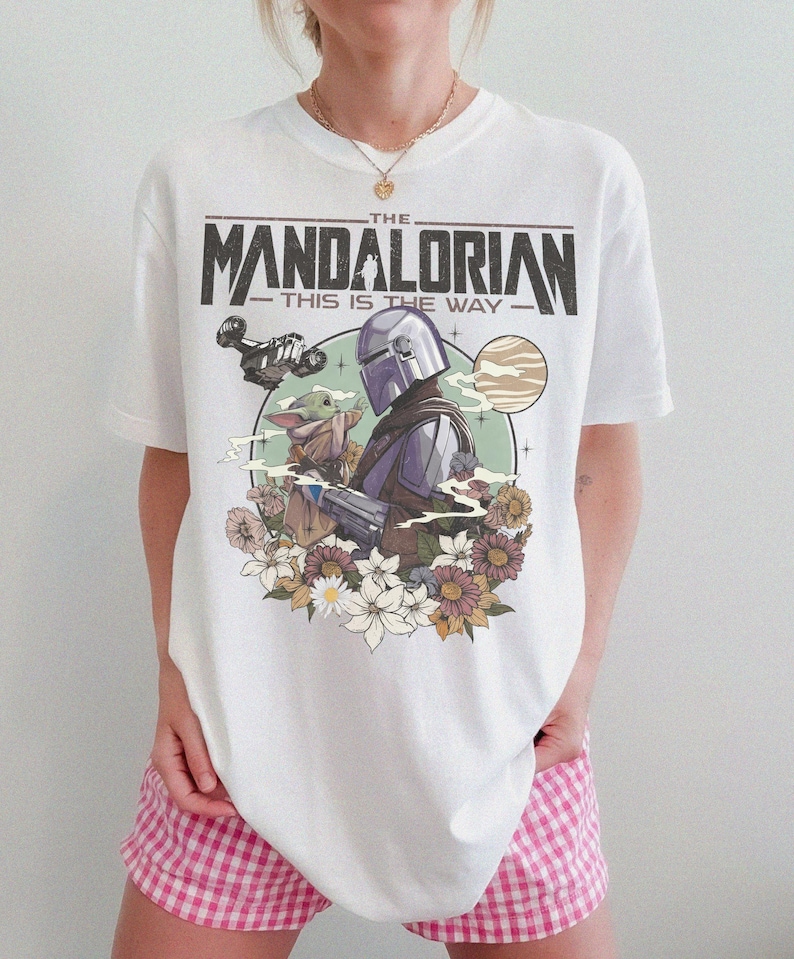 Puede incluir: Una camiseta blanca de manga corta con un estampado gr&aacute;fico de The Mandalorian y Grogu. El dise&ntilde;o incluye el texto "THE MANDALORIAN" y "THIS IS THE WAY" sobre los personajes, rodeado por un fondo circular, planetas y flores.