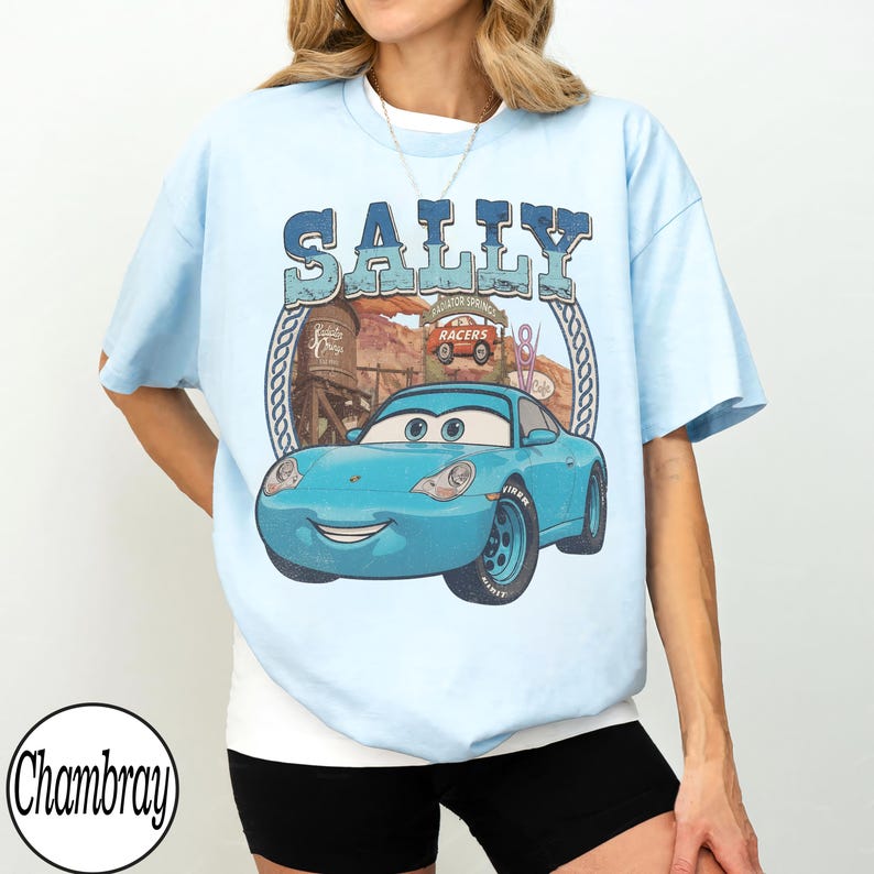 Puede incluir: Una camiseta azul claro con un gr&aacute;fico del personaje Sally de la pel&iacute;cula Cars. El dise&ntilde;o incluye el texto Sally, un paisaje des&eacute;rtico, una torre de agua y un coche rojo. La prenda tiene mangas cortas y un cuello redondo.