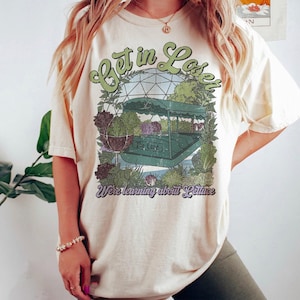 May include: Cream-colored t-shirt with a graphic featuring a green boat, plants, and the text "Get in Lose It" and "We're learning about Lettuce". The design has a vintage, retro style.