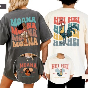 Puede incluir: Dos camisetas, una gris y otra crema, con gr&aacute;ficos. La camiseta gris presenta el texto "MOANA" y una ilustraci&oacute;n de personaje. La camiseta crema muestra el texto "HEI HEI" y una ilustraci&oacute;n de gallo. Ambas camisetas tienen texto adicional.