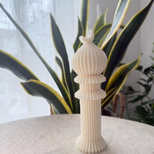Puede incluir: Vela de pilar de color marfil con un dise&ntilde;o arquitect&oacute;nico &uacute;nico. La vela presenta una base de columna acanalada, dos volantes en capas y una parte superior en forma de c&uacute;pula. La vela no est&aacute; perfumada y se encuentra sobre una superficie clara, con plantas verdes y amarillas en el fondo.