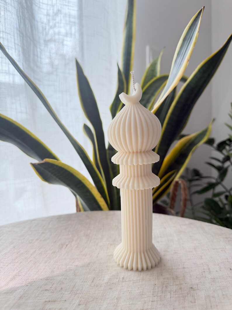 Puede incluir: Vela de pilar de color marfil con un dise&ntilde;o arquitect&oacute;nico &uacute;nico. La vela presenta una base de columna acanalada, dos volantes en capas y una parte superior en forma de c&uacute;pula. La vela no est&aacute; perfumada y se encuentra sobre una superficie clara, con plantas verdes y amarillas en el fondo.