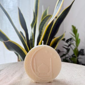 Puede incluir: Una vela redonda de color blanquecino con un dise&ntilde;o de luna creciente y mezquita. La vela tiene una mecha en la parte superior y est&aacute; colocada sobre un fondo de plantas verdes y amarillas. La vela est&aacute; sobre una superficie clara y texturizada.