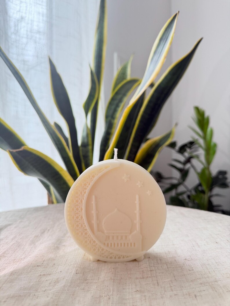 Puede incluir: Una vela redonda de color blanquecino con un dise&ntilde;o de luna creciente y mezquita. La vela tiene una mecha en la parte superior y est&aacute; colocada sobre un fondo de plantas verdes y amarillas. La vela est&aacute; sobre una superficie clara y texturizada.