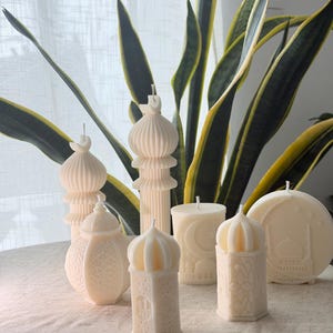Puede incluir: Un surtido de velas de color marfil en varias formas, incluyendo una mezquita, una linterna y una vela cil&iacute;ndrica con un dise&ntilde;o de luna creciente. Las velas est&aacute;n dispuestas sobre una superficie blanca con hojas de plantas verdes y amarillas en el fondo.