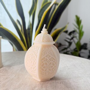 Puede incluir: Una vela decorativa de color crema con forma de linterna. La vela tiene un patr&oacute;n en relieve detallado y una peque&ntilde;a tapa abovedada. Una mecha blanca se extiende desde la parte superior. La vela est&aacute; sobre una superficie de color neutro, con plantas verdes al fondo.