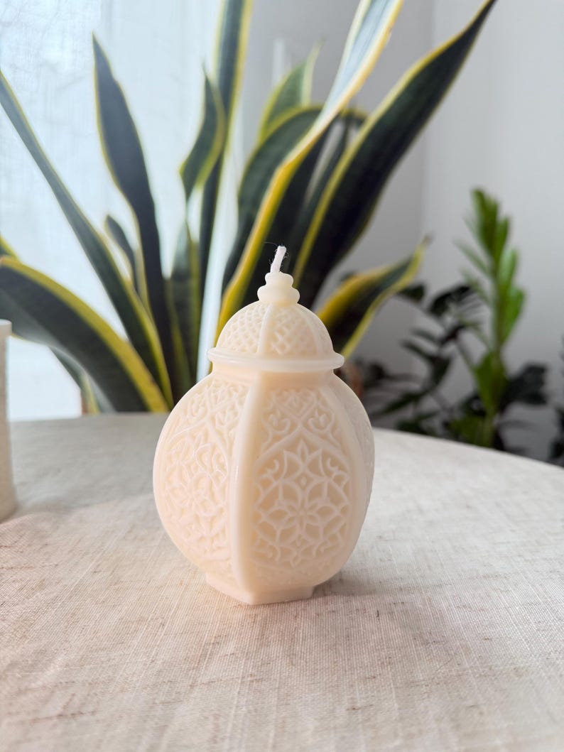 Puede incluir: Una vela decorativa de color crema con forma de linterna. La vela tiene un patr&oacute;n en relieve detallado y una peque&ntilde;a tapa abovedada. Una mecha blanca se extiende desde la parte superior. La vela est&aacute; sobre una superficie de color neutro, con plantas verdes al fondo.