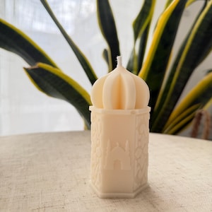 Puede incluir: Una vela de color crema con una parte superior en forma de c&uacute;pula y una base hexagonal. La vela presenta un dise&ntilde;o detallado de una mezquita y motivos florales. La vela est&aacute; colocada sobre una superficie texturizada de color claro, con follaje verde y amarillo en el fondo.