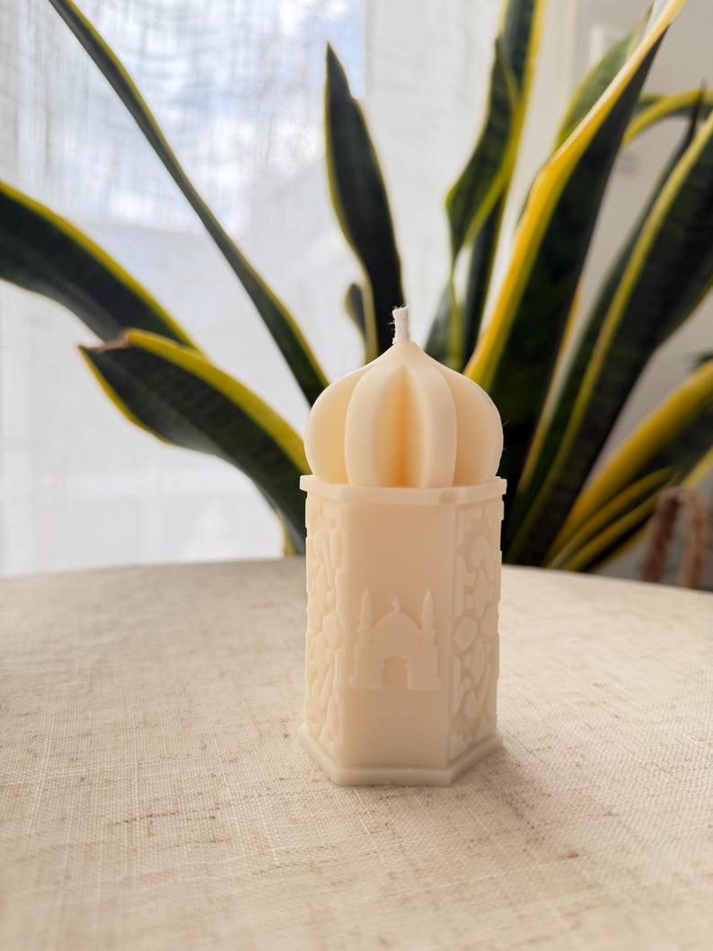 Puede incluir: Una vela de color crema con una parte superior en forma de c&uacute;pula y una base hexagonal. La vela presenta un dise&ntilde;o detallado de una mezquita y motivos florales. La vela est&aacute; colocada sobre una superficie texturizada de color claro, con follaje verde y amarillo en el fondo.