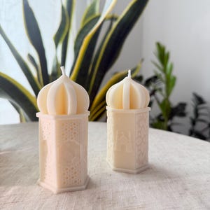 Puede incluir: Dos velas decorativas de color crema con forma de faroles ornamentados. Las velas tienen un patr&oacute;n geom&eacute;trico detallado en la base y una parte superior en forma de c&uacute;pula. Las velas est&aacute;n sobre una superficie de color neutro, con plantas verdes en el fondo.
