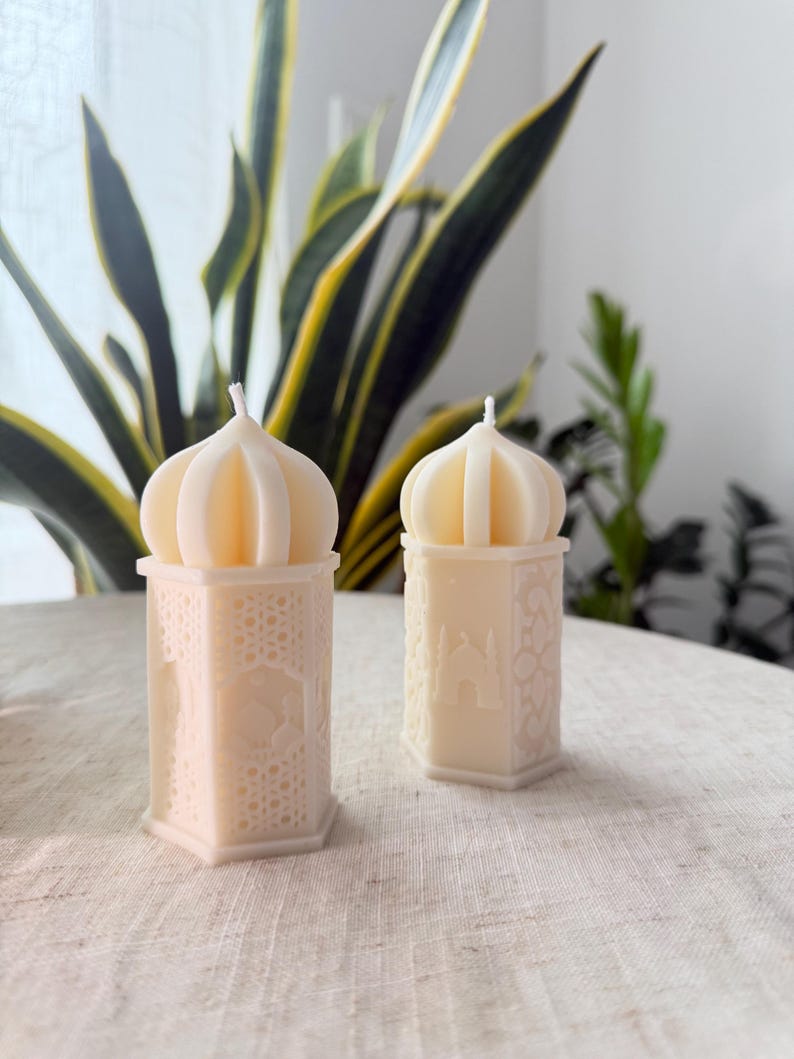 Puede incluir: Dos velas decorativas de color crema con forma de faroles ornamentados. Las velas tienen un patr&oacute;n geom&eacute;trico detallado en la base y una parte superior en forma de c&uacute;pula. Las velas est&aacute;n sobre una superficie de color neutro, con plantas verdes en el fondo.