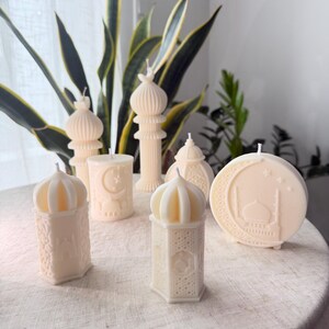 Puede incluir: Un surtido de velas de color marfil en varias formas, incluyendo faroles, minaretes y una vela redonda con un dise&ntilde;o de mezquita. Las velas est&aacute;n dispuestas sobre una superficie de tono neutro, con vegetaci&oacute;n en el fondo.