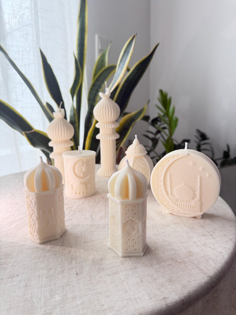 Puede incluir: Un surtido de velas de color marfil en varias formas, incluyendo faroles, minaretes y una vela redonda con un dise&ntilde;o de mezquita. Las velas est&aacute;n dispuestas sobre una superficie de tono neutro, con vegetaci&oacute;n en el fondo.