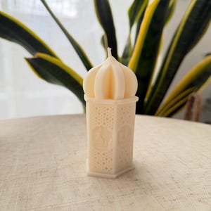 Puede incluir: Vela de color marfil con forma de linterna y parte superior abovedada. La vela tiene intrincados patrones recortados y un dise&ntilde;o detallado de un edificio en cada lado. La vela est&aacute; colocada sobre una superficie de color neutro, con una planta en el fondo.