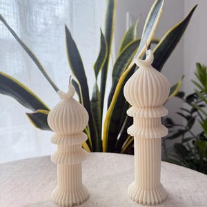 Puede incluir: Dos velas de pilar de color marfil con un dise&ntilde;o &uacute;nico. Cada vela presenta un cuerpo acanalado en capas y un detalle de media luna en la parte superior. Las velas se colocan sobre una superficie de color claro, con plantas verdes en el fondo.