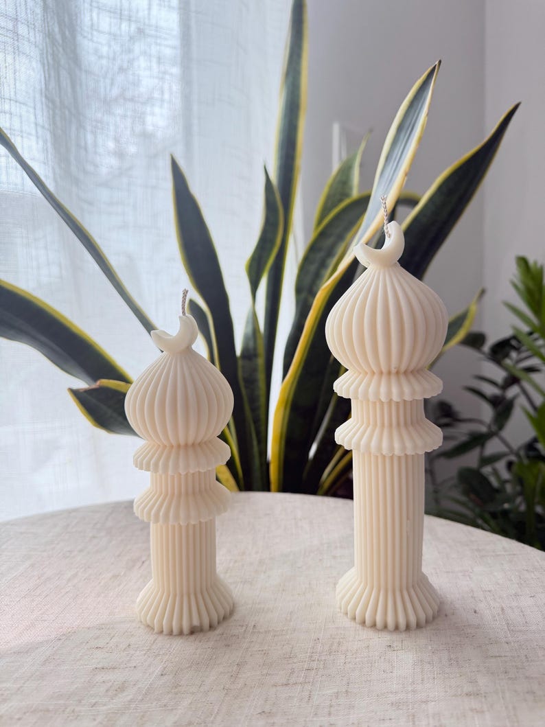 Puede incluir: Dos velas de pilar de color marfil con un dise&ntilde;o &uacute;nico. Cada vela presenta un cuerpo acanalado en capas y un detalle de media luna en la parte superior. Las velas se colocan sobre una superficie de color claro, con plantas verdes en el fondo.