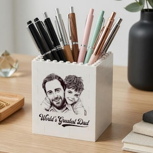 Puede incluir: Un portalápices blanco con una imagen en blanco y negro de un padre y un niño, y el texto "World's Greatest Dad". El portalápices contiene varios bolígrafos en negro, plateado, rosa y veteado de madera. También se ve una bandeja de madera con clips y un calendario.